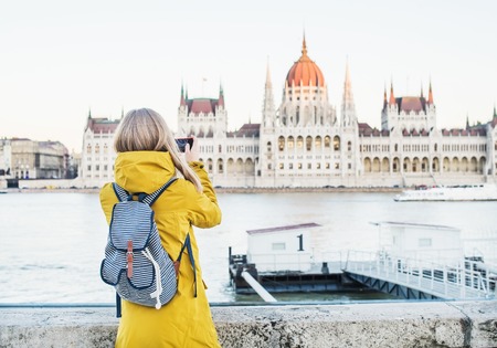 Young blondy woman tourist making photos of Parliament historic building from Danube side with her phone in Budapest, Hungary, on a spring clear dayの写真素材