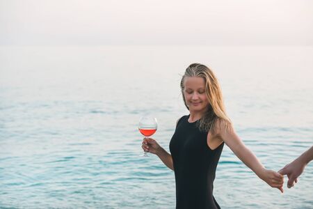 Young blond woman enjoying glass of rose wine and holding man's hand on beach by the sea at sunset....
