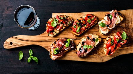 Brushetta set for wine. Variety of small sandwiches with prosciutto, tomatoes, parmesan cheese, fresh basil and balsamic creme served with glass of red wine on rustic wooden board over dark background, top viewの写真素材