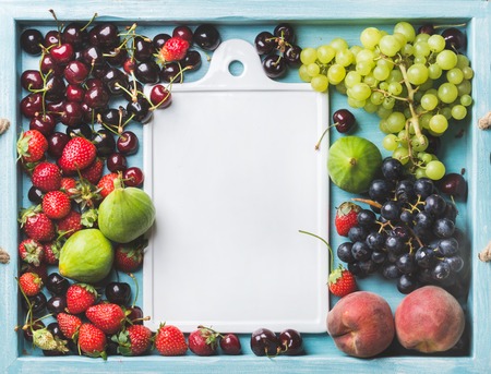 Healthy summer fruit variety. Figs, black and green grapes, sweet cherries, strawberries and peaches on blue painted wooden background with white ceramic board in center, top view, copy spaceの写真素材