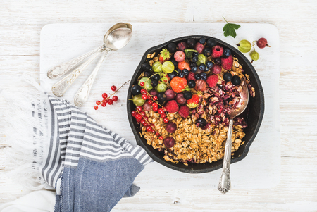 Healthy breakfast. Oat granola crumble with fresh berries and seeds in iron skillet pan on wooden board over white wooden backdrop, top view, horizontal compositionの写真素材