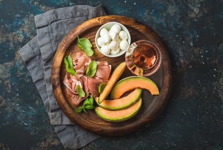 Italian antipasti snack for wine. Prosciutto ham, cantaloupe melon, mozzarella cheese, fresh basil leaves and glass of rose on wooden round serving board over grunge dark plywood background,top viewの写真素材