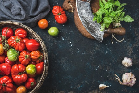 Fresh colorful ripe heirloom tomatoes in basket, basil leaves, garlic, herb chopper knife for cooking over grunge dark plywood background. Top view, copy space. Harvest vegetable cooking conception.の写真素材