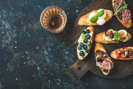 Italian crostini with various toppings on round wooden board and glass of rose wine over black plywood background, top view, copy space, horizontal compositionの写真素材