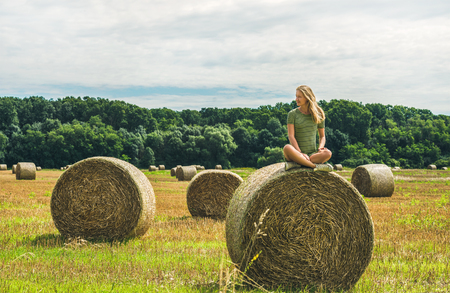 Young blond girl with long hair sitting legs crossed on haystack and smiling with closed eyes on sunny summer day, Badasconytomaj, Hungaryの写真素材