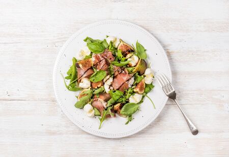 Prosciutto, arugula, basil, figs and mozzarella salad over white wooden background, top viewの写真素材