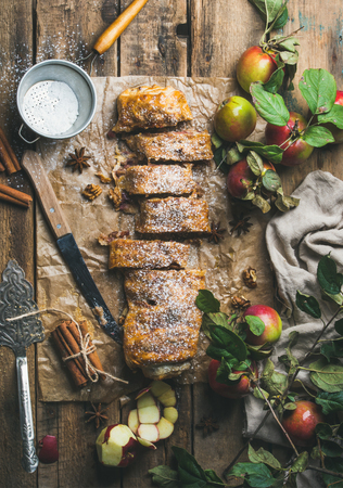 Apple strudel cake with cinnamon and sugar powder cut in slices served with star anise, nuts and fresh apples on rustic wooden background, top view, vertical compositionの写真素材