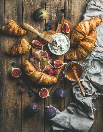 Breakfast with freshly baked croissants, ricotta cheese, figs, fresh berries, prosciutto di Parma and honey in glass jar over rustic wooden background, top view, vertical compositionの写真素材