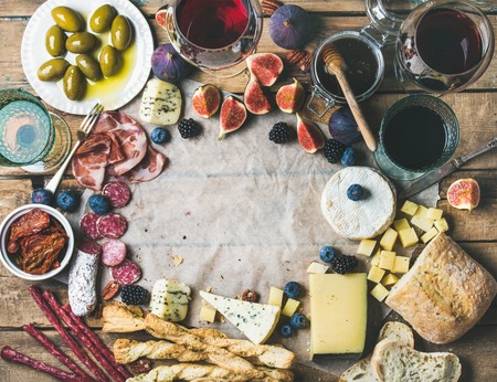 Food frame. Wine and snack set with various wines in glasses, meat variety, bread, green olives, figs and berries on wax paper over rustic wooden background, top view, copy space in centerの写真素材