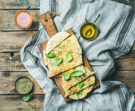 Homemade Italian focaccia flatbread cut into pieces with herbs, fresh basil leaves, olive oil and glass of rose wine on rustic serving board over wooden background, top viewの写真素材