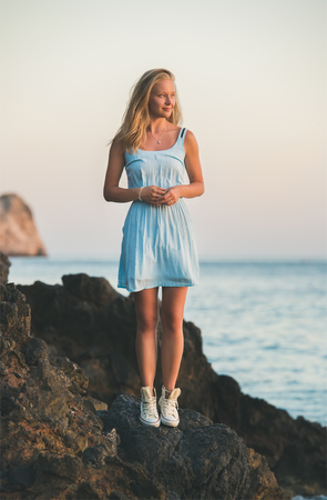 Young blond tourist woman in blue dress standing on rocks by the sea at sunset, looking at sun and smiling. Kleopatra beach, Alanya, Mediterranean region, Turkeyの写真素材
