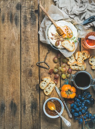 Homemade camembert cheese in small pan, honey, fig jam, persimmon, grapes, pecan nuts, grilled baguette slices and glass of rose wine over rustic wooden background, top view, copy spaceの写真素材