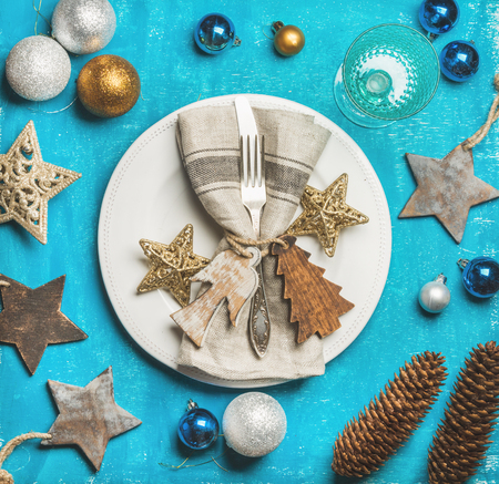 Christmas, New Year holiday table setting. Plate with linen napkin, napkin holders, pine cones, silverware and golden stars, wine glass over bright blue background. Party, family celebration conceptの写真素材