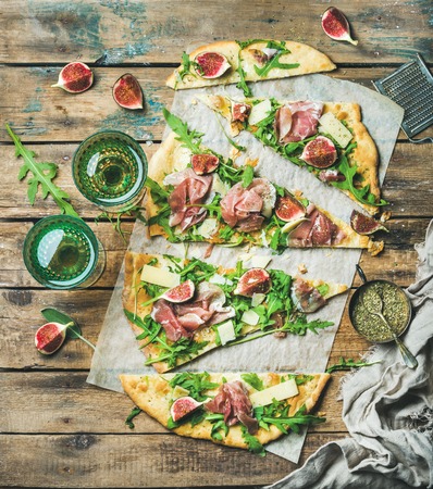 Fig, prosciutto, arugula and sage flatbread pizza cut into pieces with glasses of rose wine over rustic wooden background, top viewの写真素材