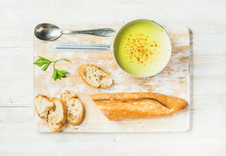 Green vegetable soup in scoop with handle with parsley and baguette on white shabby wooden board over white painted wooden background, top view, horizontal compositionの写真素材