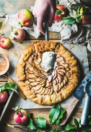 Man's hand holding piece of homemade apple crostata with cinnamon and ice-cream scoop over whole pie served with fresh garden apples with leaves on rustic wooden background, top view, selective focusの写真素材
