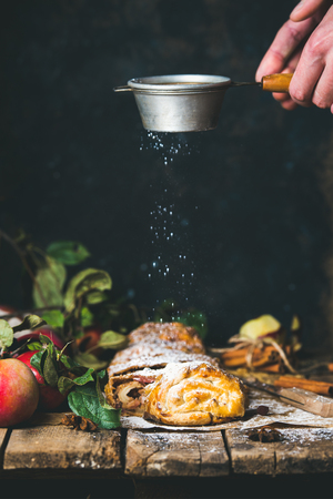 Apple strudel cake cut in pieces with fresh red apples on rustic wooden table and man's hands with sieve sprinkling sugar powder from above, dark plywood wall background, vertical compositionの写真素材