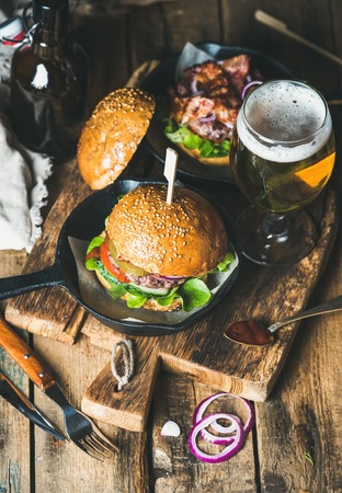 Homemade beef burgers with crispy bacon and vegetables in small black pan and glass of wheat beer on rustic serving board over shabby wooden background, top view, selective focus, vertical compositionの写真素材