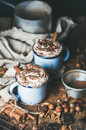 Hot chocolate in blue mugs with whipped cream and cinnamon sticks, spices, nuts and cocoa powder on rustic wooden background, selective focus, vertical compositionの写真素材