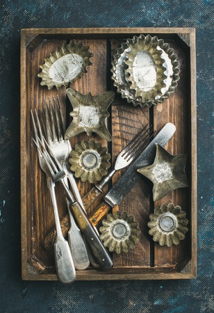 Old vintage tin baking molds and cutlery in rustic wooden tray over dark blue shabby background, top viewの写真素材