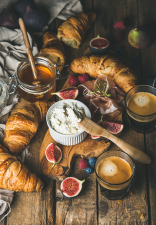 Breakfast with croissants, ricotta cheese, figs, fresh berries, prosciutto meat, honey in glass jar and espresso coffee over rustic wooden background, selective focus, vertical compositionの写真素材