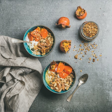 Healthy vegetarian breakfast. Oatmeal, quinoa granola with yogurt, dried fruit, seeds, honey, persimmon in bowls over grey background, top view, square crop. Gluten free, allergy-friendly conceptの写真素材