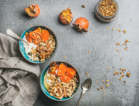 Healthy vegetarian breakfast. Oatmeal, quinoa granola with yogurt, dried fruit, seeds, honey, persimmon in bowls over grey background, top view, copy space. Gluten free, allergy-friendly conceptの写真素材