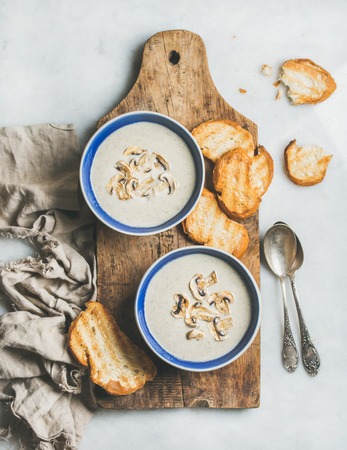 Creamy mushroom soup in bowls with toasted bread slices on rustic serving board over grey marble background, top view, selective focusの写真素材