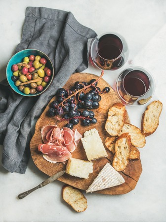 Wine and snack set. Variety of cheese, olives in blue bowl, prosciutto, roasted baguette slices, black grapes on wooden board and glasses of red wine over grey marble background, top viewの写真素材
