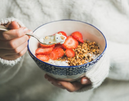 Healthy breakfast greek yogurt, granola and strawberry bowl in hands of woman wearing white loose knitted woolen sweater, selective focus. Clean eating, healthy, vegetarian, dieting food conceptの写真素材