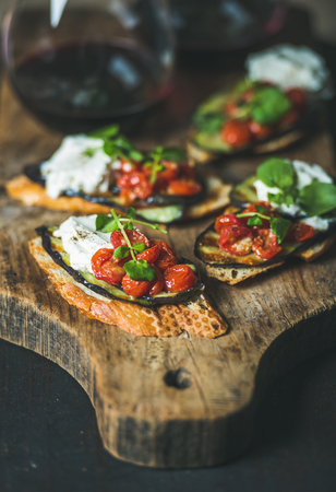 Wine and snack set. Brushetta with roasted eggplant, cherry tomatoes, garlic, cream cheese, arugula and glass of red wine on wooden board over dark background, selective focus. Slow food conceptの写真素材