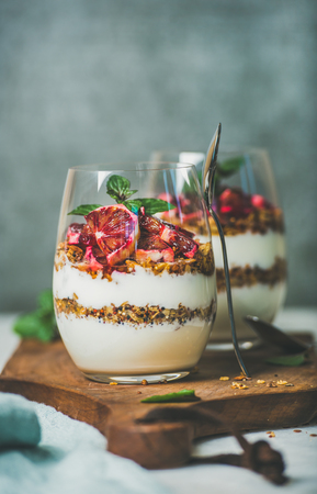 Healthy breakfast. Greek yogurt, granola, blood orange layered parfait in glasses with fresh mint on wooden board, grey concrete wall at background, copy space. Clean eating, detox food conceptの写真素材