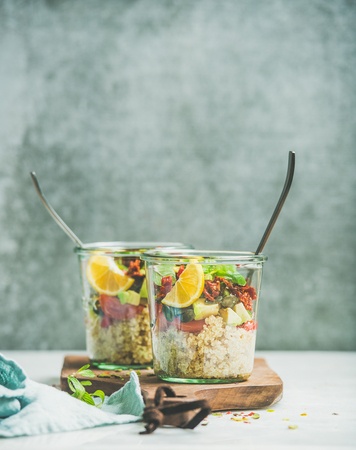 Healthy vegetarian salad with quionoa, avocado, dried tomatoes, basil, mint leaves, orange in glass jars on wooden board, grey wall at background, selective focus, copy space. Weight loss food conceptの写真素材