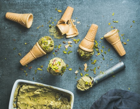 Homemade pistachio ice cream in ceramic mold and metal scooper with crashed pistachio nuts and waffle cones over grey concrete background, top view, selective focusの写真素材
