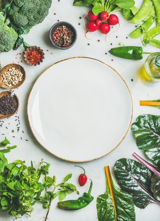 Fresh raw greens, unprocessed vegetables and grains over light grey marble kitchen countertop, wtite plate in center, top view, copy space. Healthy, clean eating, vegan, detox, dieting food conceptの写真素材