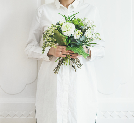 Young woman wearing white clothes holding white flowers bouquet composed of ranunculus and lilacs in her hands, llight wall at background. Wedding, flower shop conceptの写真素材