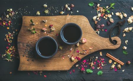 Chinese black tea in black stoneware cups on serving wooden board over black wooden background with herbs, flower buds, tea leaves spilt around, top viewの写真素材