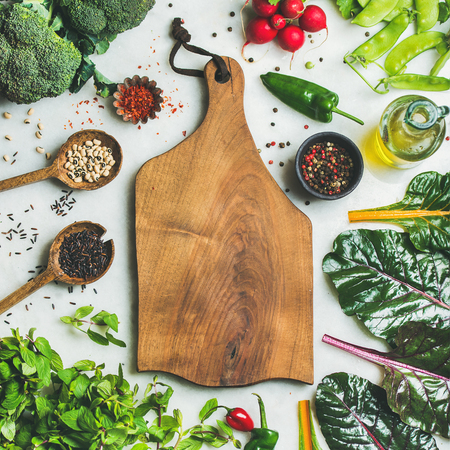 Fresh raw greens, unprocessed vegetables and grains over light grey marble kitchen countertop, wooden board in center, top view, copy space, square crop. Healthy, vegan, detox, dieting food conceptの写真素材
