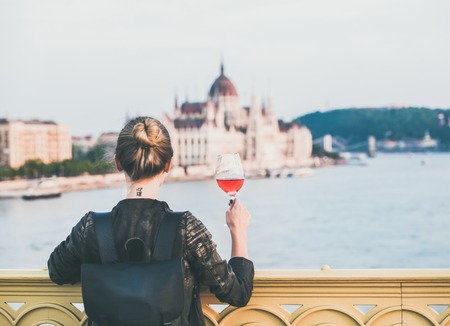 Young woman tourist standing with glass of rose wine at Margaret bridge in Budapest, Hungarian Parliament building and Duna river at backgroundの写真素材
