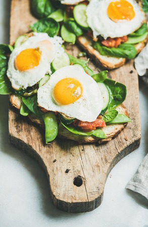Healthy breakfast sandwiches. Bread toasts with fried eggs and fresh vegetables on rustic wooden board over grey marble background, selective focus. Clean eating, healthy, weight loss food conceptの写真素材