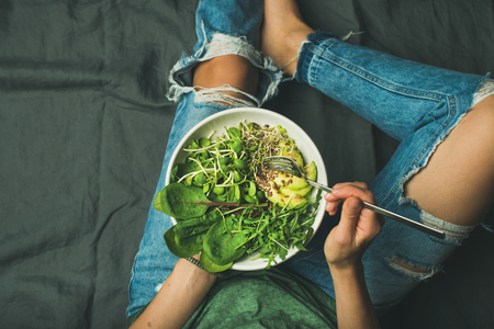 Green vegan breakfast meal in bowl with spinach, arugula, avocado, seeds and sprouts. Girl in jeans holding fork with knees and hands visible, top view, copy space. Clean eating, vegan food conceptの写真素材