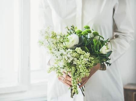 Young woman wearing white clothes holding white flowers bouquet composed of ranunculus and lilacs in her hands beside window. Wedding, flower shop conceptの写真素材