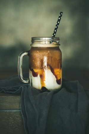 Iced caramel macciato coffee with milk in glass jar with straw on dark rustic wooden table, selective focus. Cold sweet refreshing summer drink conceptの写真素材