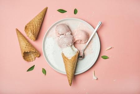 Flatlay of pastel pink strawberry and coconut ice cream scoops, sweet waffle cones on white plate...