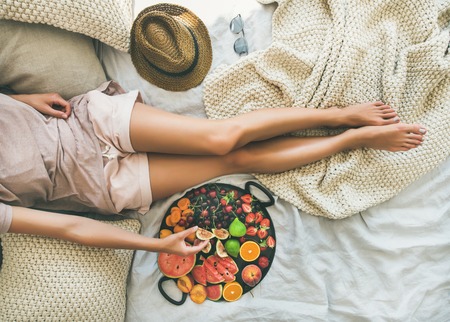 Summer healthy raw vegan clean eating breakfast in bed concept. Young girl wearing pastel colored home clothes taking fruit from tray full of fresh seasonal fruit. Top view, horizontal compositionの写真素材