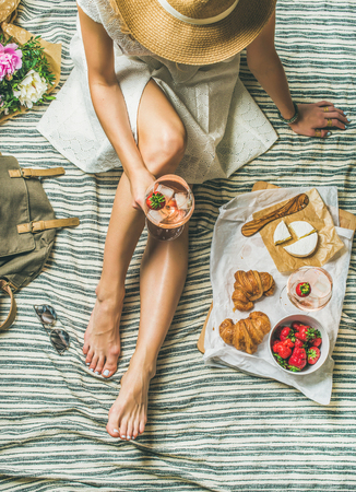 French style romantic picnic setting. Young woman in dress with glass of rose wine, fresh strawberries, croissants, brie cheese, sunglasses, peony flowers, top view. Outdoor gathering conceptの写真素材