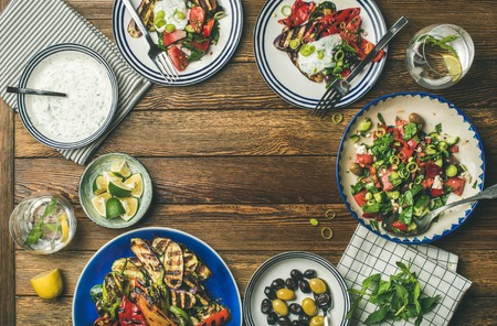 Flat-lay of healthy dinner table setting. Fresh salad, grilled vegetables with yogurt sauce, pickled olives over wooden background, copy space, selective focus. Clean eating, vegan, vegetarian foodの写真素材