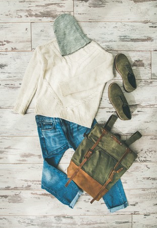 Flat-lay of Fall lady' s clothes outfit. Ecru colored knitted woolen sweater with braids, blue denim jeans, haki suede oxford boots, backpack, cap over parquet background, top view. Trendy Autumn lookの写真素材