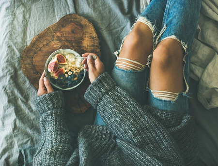 Healthy winter breakfast in bed. Woman in grey sweater and jeans eating rice coconut porridge with figs, berries, hazelnuts, top view. Clean eating, diet, vegetarian, vegan, comfort food conceptの写真素材