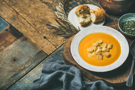 Fall warming pumpkin cream soup with croutons and seeds on board over rustic wooden background, copy space, selective focus. Autumn vegetarian, vegan, healthy comfort food conceptの写真素材